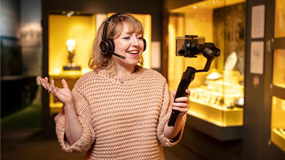 Kvinna med hörlurar håller och tittar in i en mobiltelefon och ler