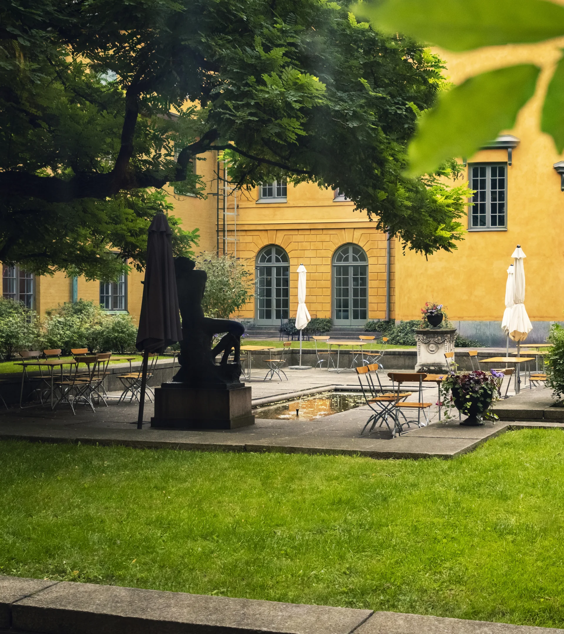 Kafébord står utställda runt en fontän i mitten av museets innergård, omgivna av grönskande träd och gräs.