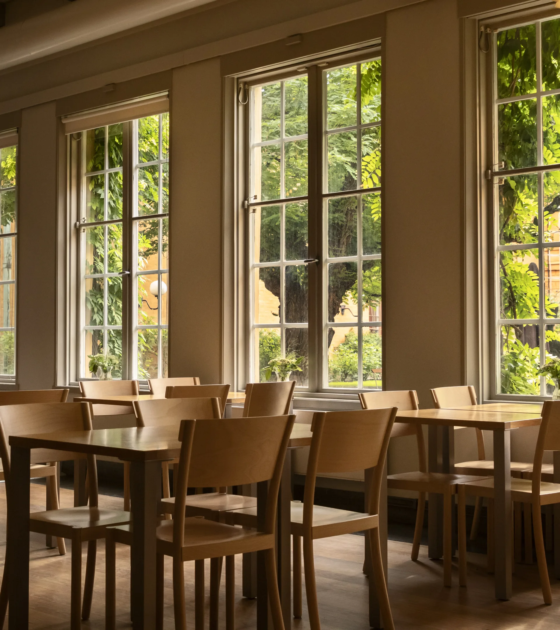 Bord med stolar framför höga fönster. Utanför syns grönskande träd.