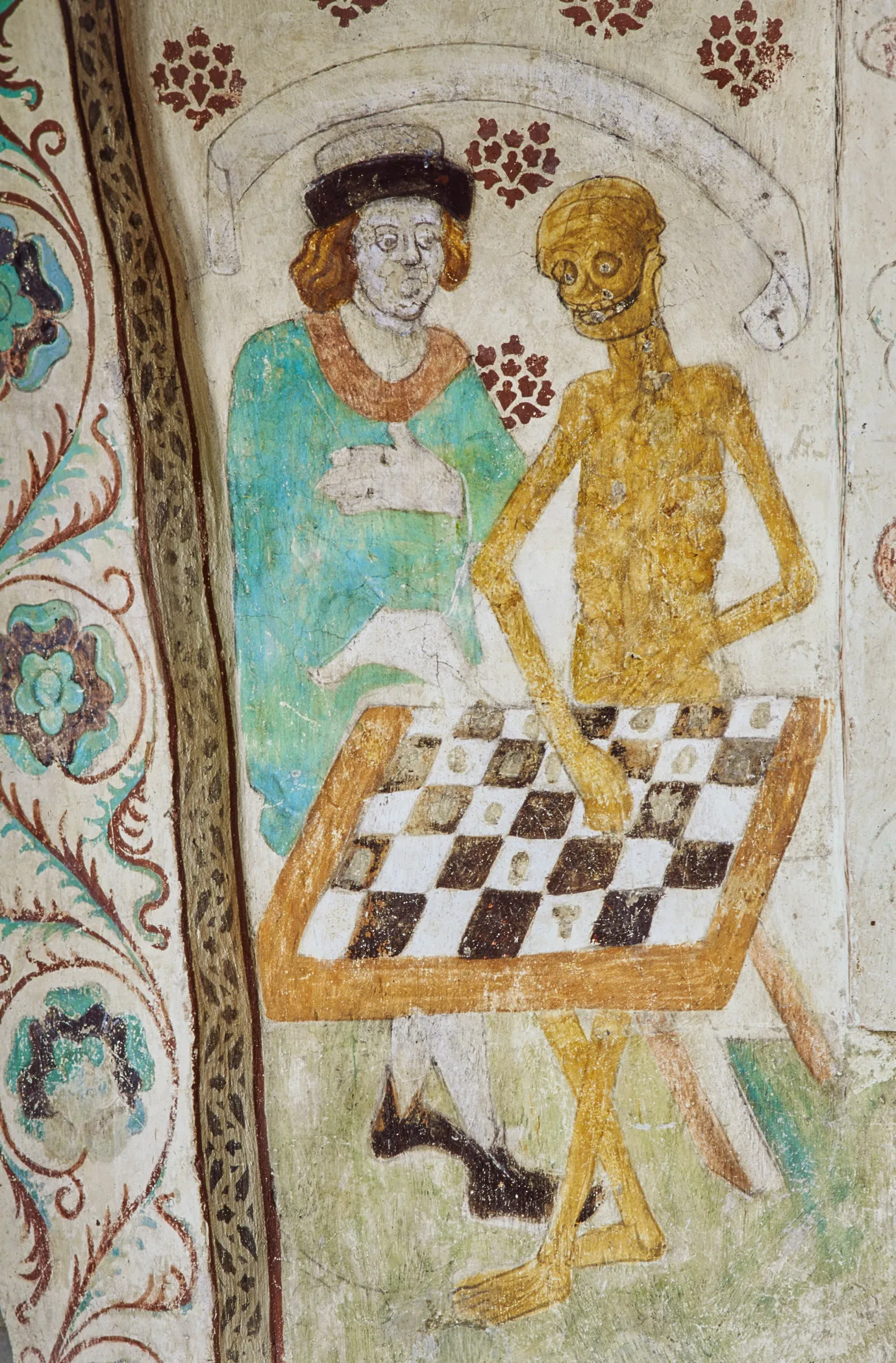 A long-haired man in green tunic and brown cap playing chess with a corpse (death).