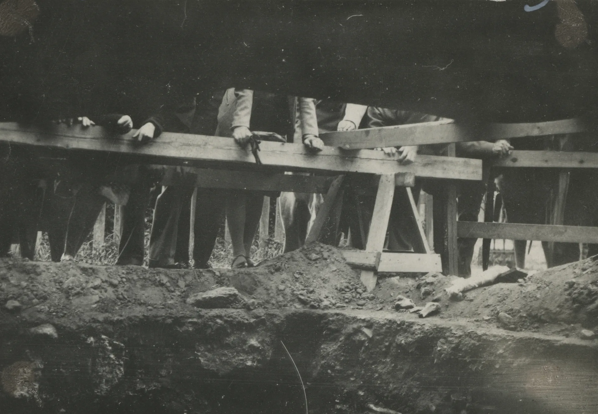 Svartvit bild som visar en utgrävningsplats. Flera personer står bakom ett trästaket vid utgrävningens kant, endast ben och händer syns.