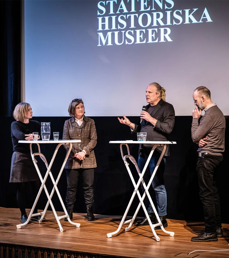 Fyra personer står på en scen vid runda höga bord. En person håller i en mikrofon.