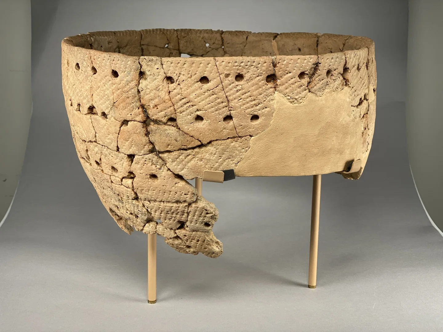 A light brown clay vessel, decorated with several rows of holes and stripes.