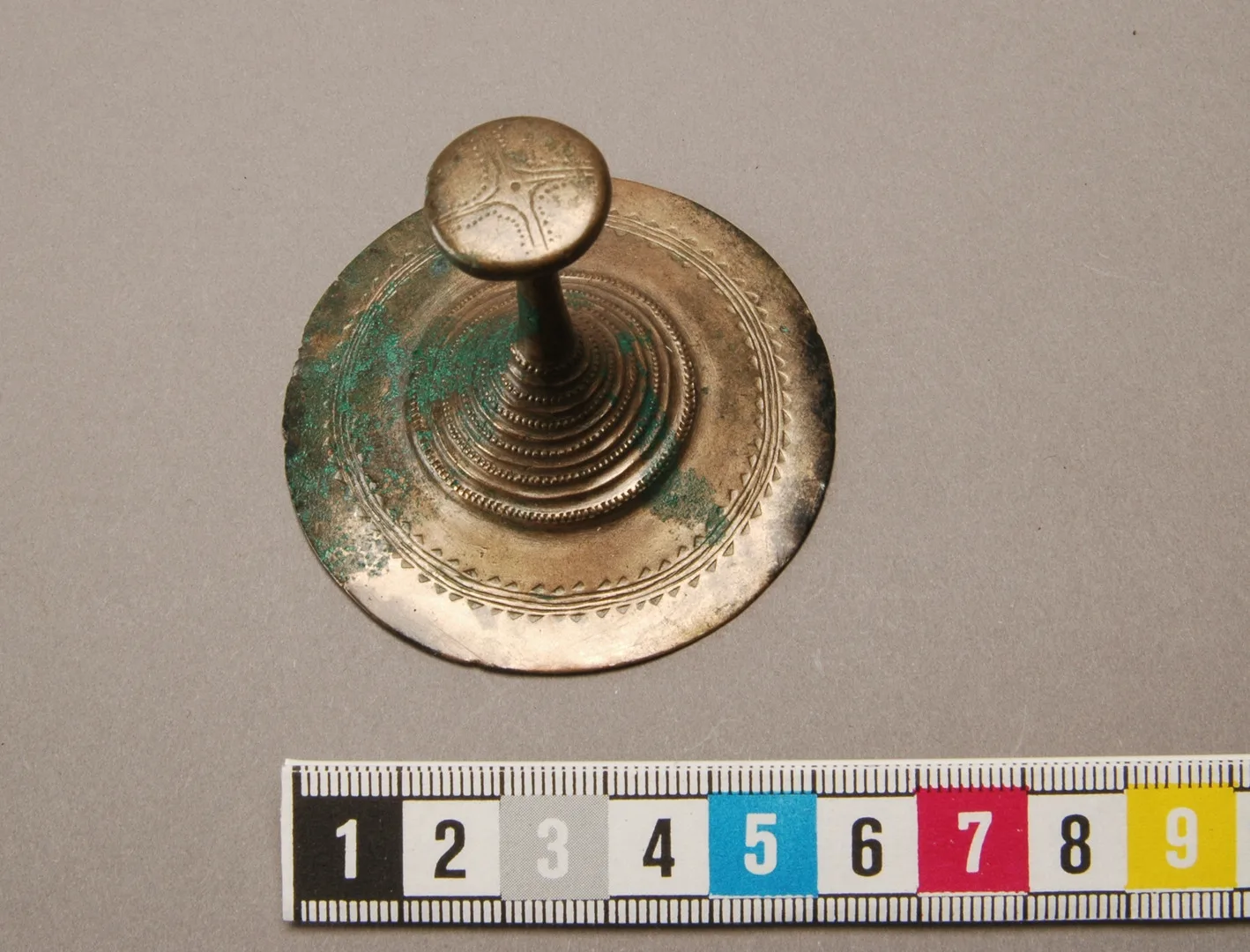 Cirkelformat föremål av kopparlegering där det sticker upp en spets med knapp