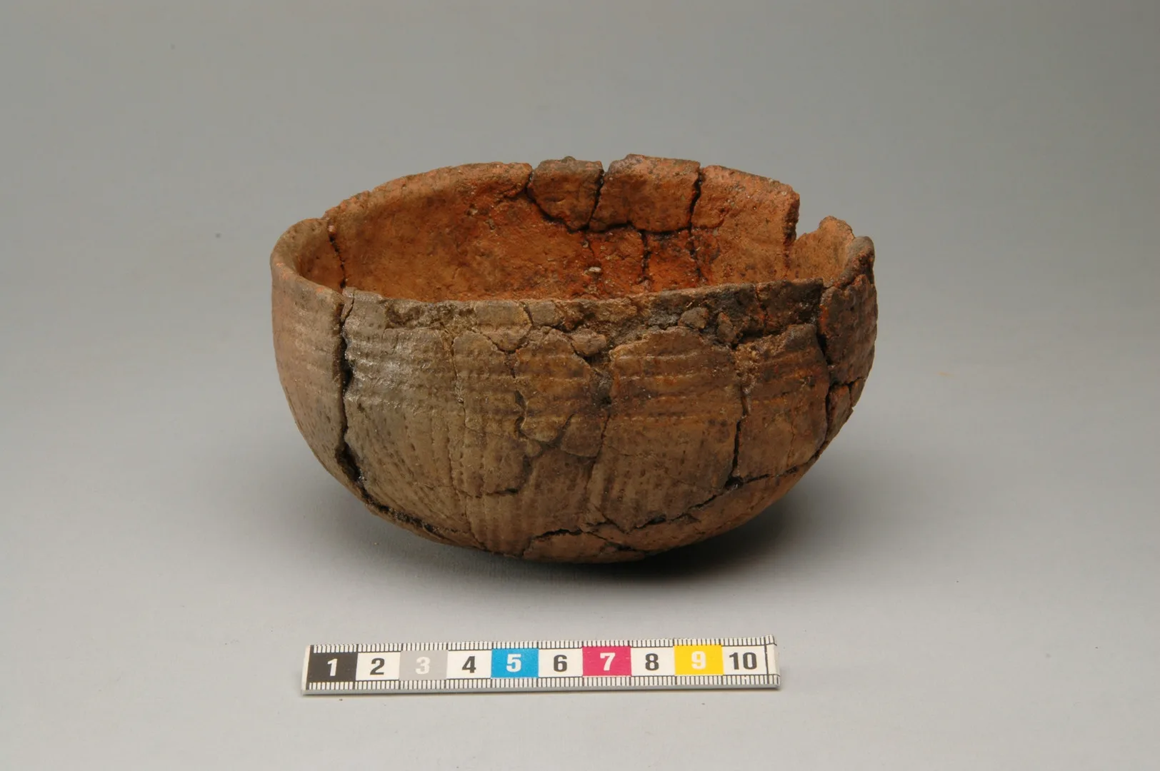 A reddish clay vessel, shaped like a hemisphere, decorated with lines.