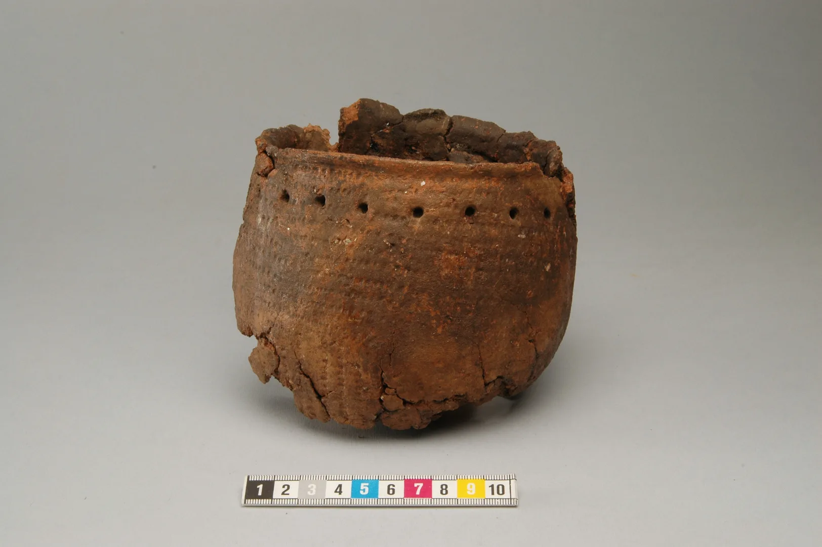A reddish clay vessel with a rounded base, decorated with dot patterns and perforations..
