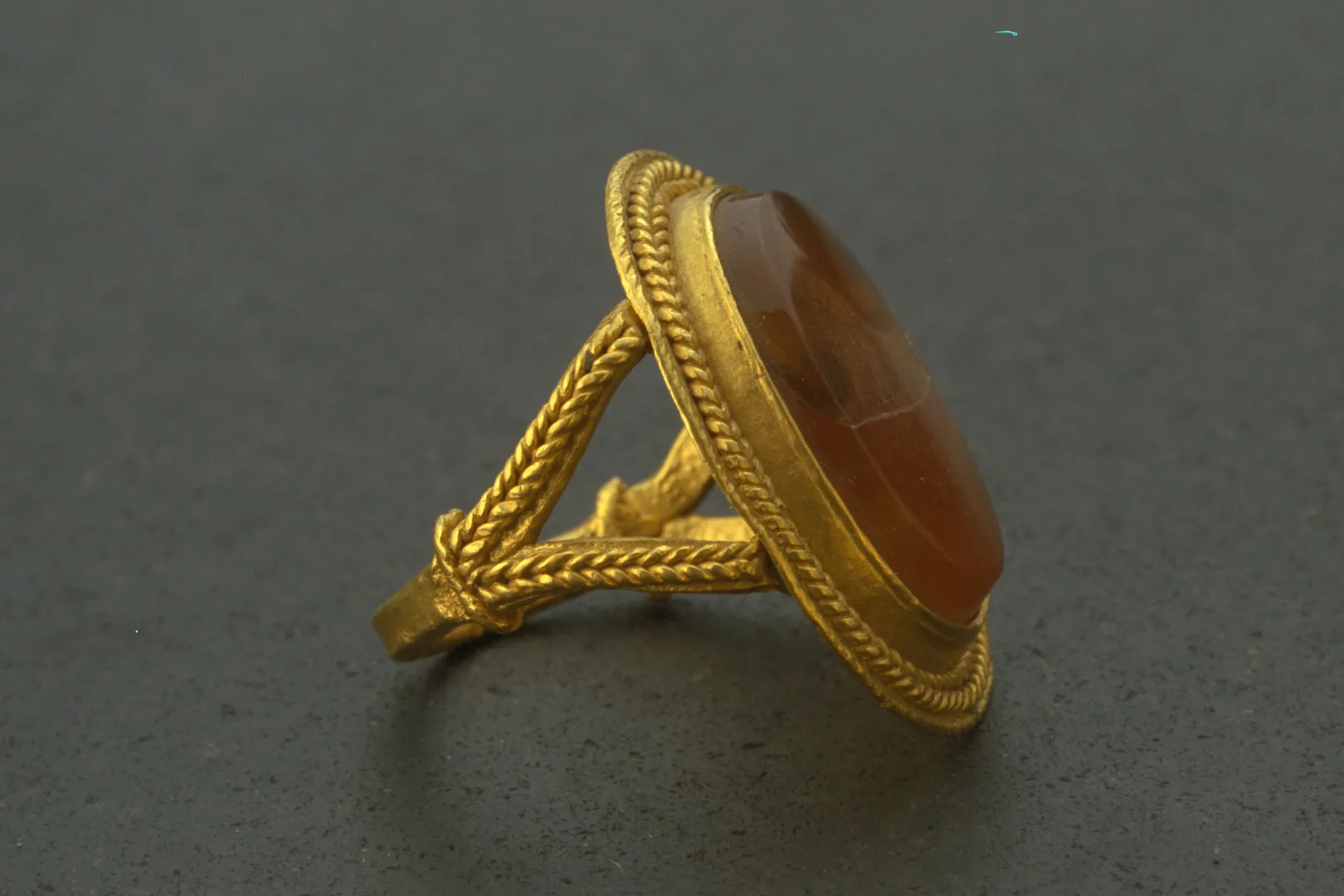 A gold ring with y-shaped supports holding an oval frame with a flat, red stone.