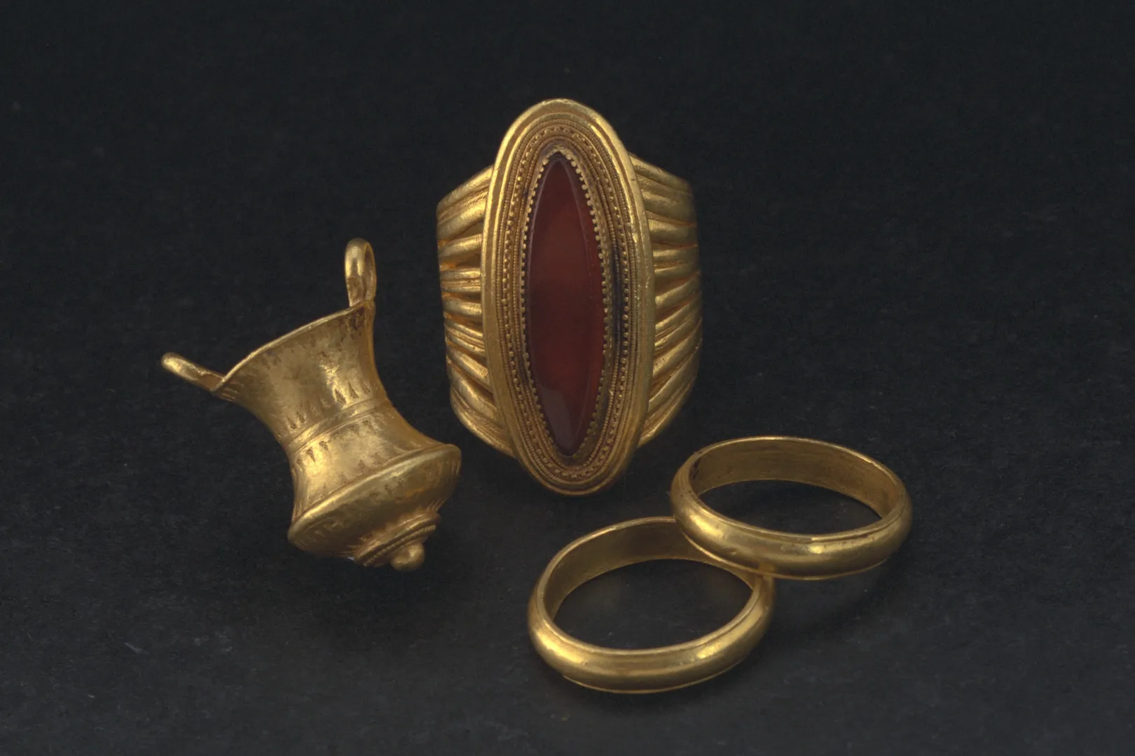 Two simple golden rings, a big golden ring with an oval red gemstone and a small golden item resembling a cauldron