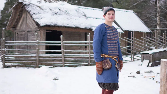 Person in reconstructed Viking Age clothes stands in front of a wooden house in the snow