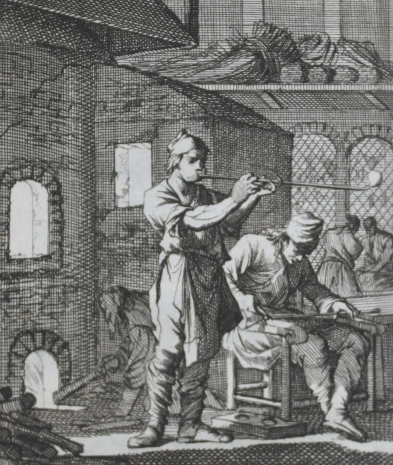 Etching in black and white depicting a man blowing into a long rod with a small piece of glass in the other end. In the background other people employed with glass making work.