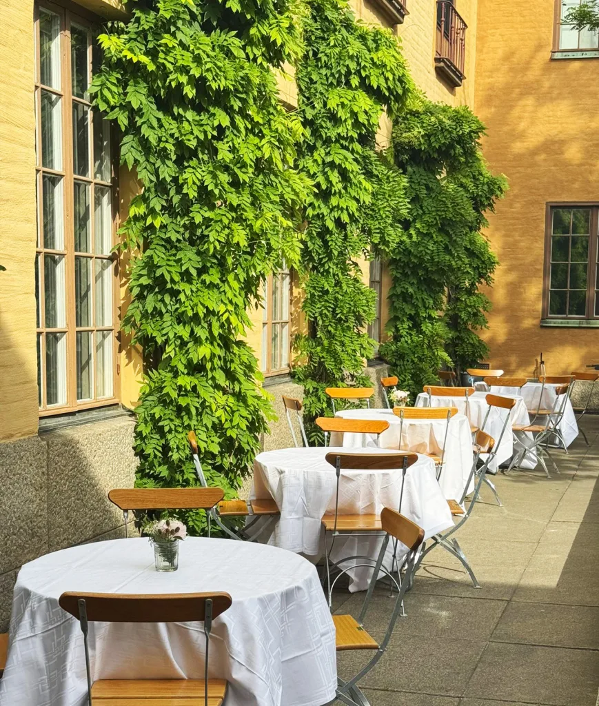 Runda mindre bord med vita dukar står utställda på museets innergård, med museets fasad i bakgrunden. På väggen slingrar sig gröna växter.