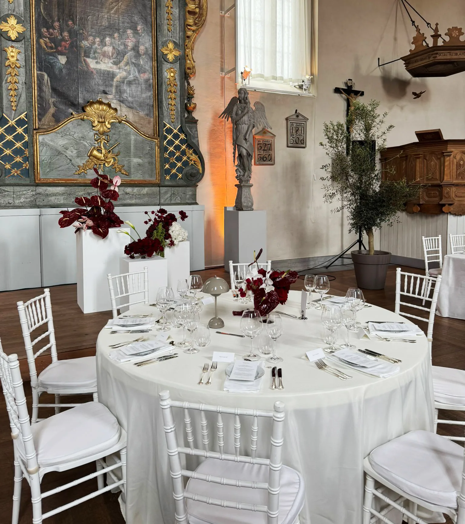 Runt dukat bord. Vit duk och vita stolar. I bakgrunden syns blomarrangermang och kyrklig konst.