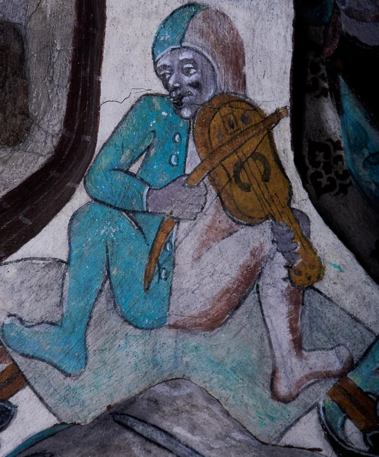 A man dressed in a two-coloured onesie sitting with his legs spread, playing a violin.