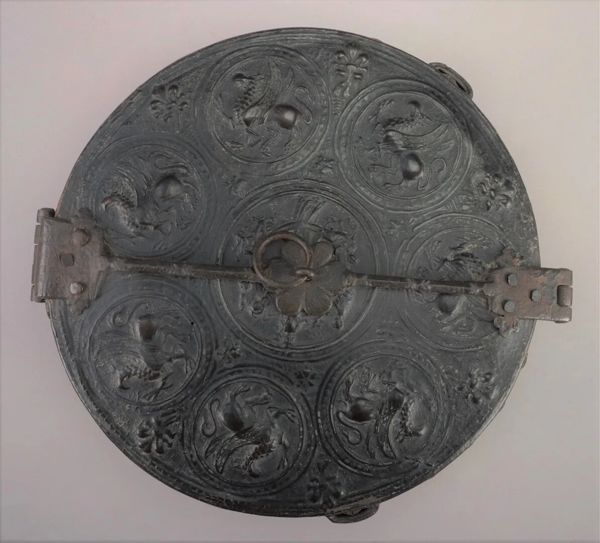 Top side of a circular leather case. Across the lid there is a metal rod with one hinge and one clasp.