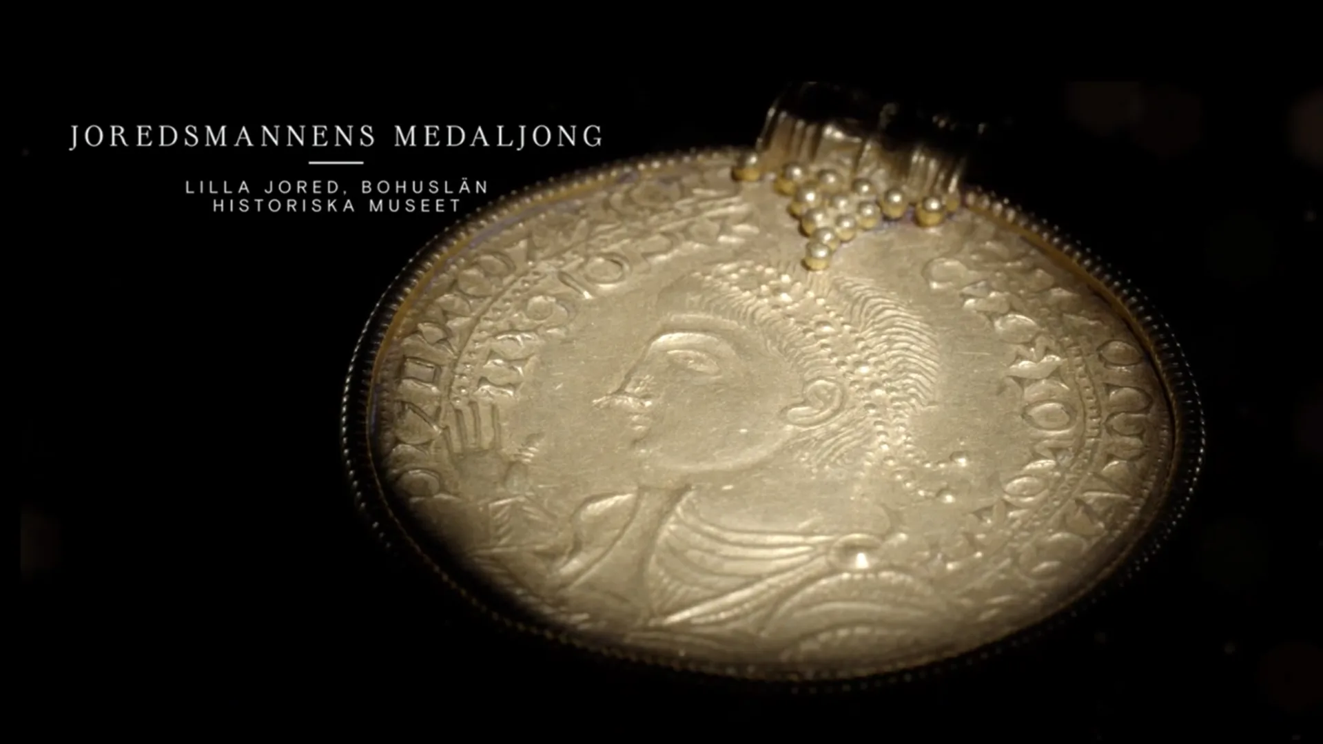 En rund medaljong. I mitten syns ett ansikte i profil som håller upp en hand.