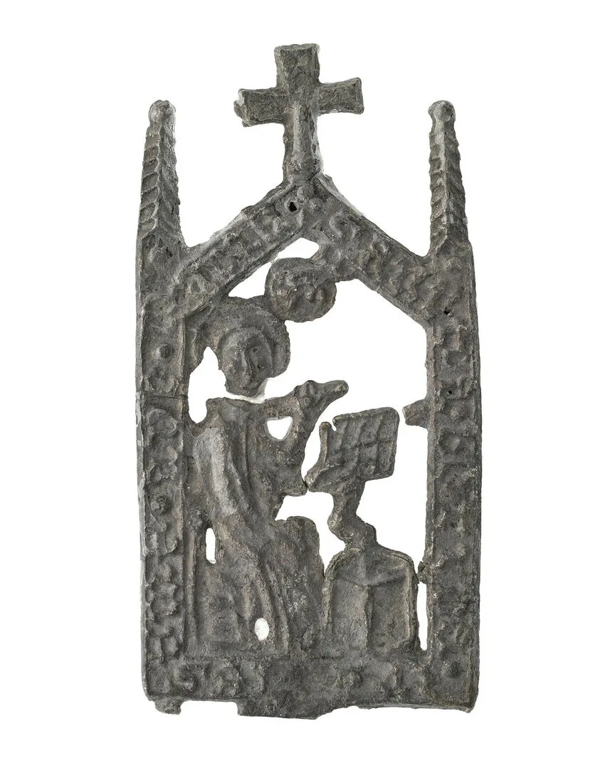 Platt metallföremål med en figur inuti en husformat ram med ett kors upptill.