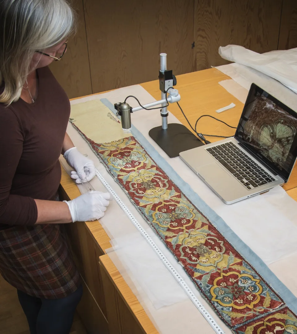 Woman with white gloves and microscope looling at colourful textile.