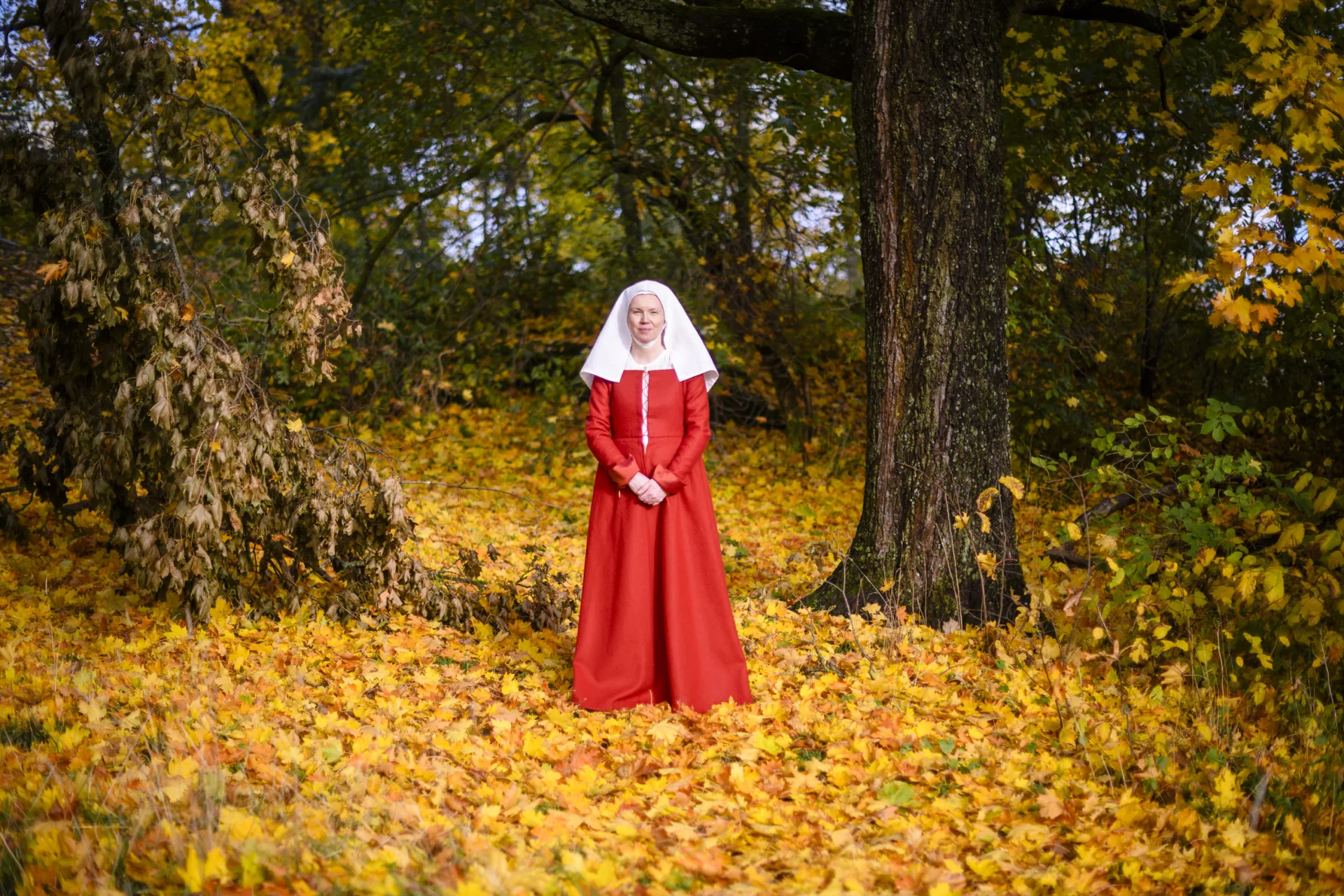 Kvinna i röd medeltida dräkt ståendes på gula löv i ett skogsparti