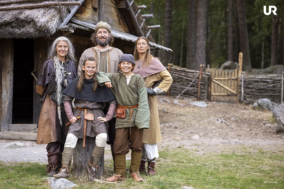 Fem personer i vikingatida kläder står framför ett hus med halmtak
